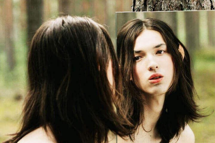 Foto do rosto de um adolescente branco, com cabelos longos, refletida em um espelho pendurado no tronco de uma árvore. A foto ilustra o artigo 10 Melhores Maneiras como a Psicanálise Impulsiona a Autoidentidade escrito pelo psicanalista Homero Mônaco para o site Online Psicanalista.