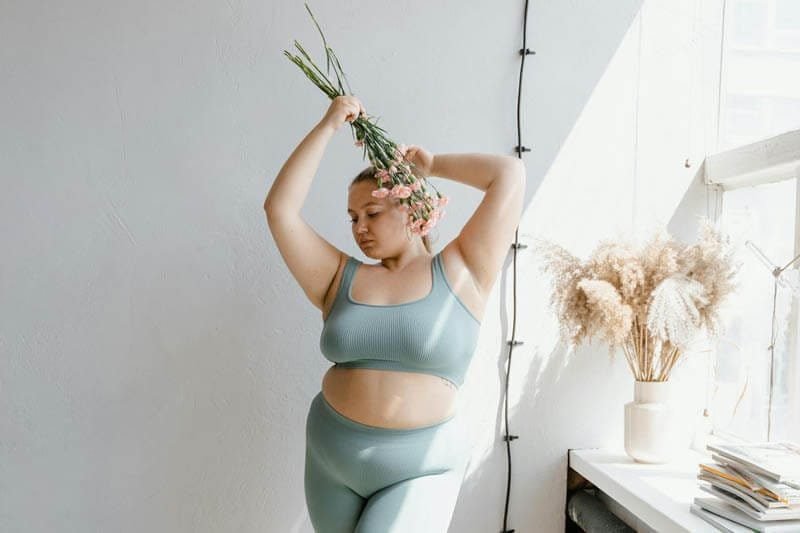 Foto de uma mulher em pé corpulenta, comm top e calça leggin cinza, e com braços para cima segurando um ramo de flores de forma que as flores fiquem próximas a sua orelha esquera. A mulher parece se sentir bem com a sua autoconfiança e a sua autoaceitação. A foto ilustra o artigo 5 Melhores Técnicas para Aumentar a sua Autoconfiança e a sua Autoaceitação escrito pelo psicanalista Homero Mônaco para o site Online Psicanalista.